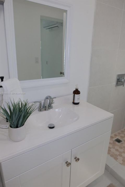 White vanity with a rectangular countertop, integrated basin, chrome faucet, and framed wall mirror, complemented by a walk-in shower with large-format wall tile and hexagonal floor tile