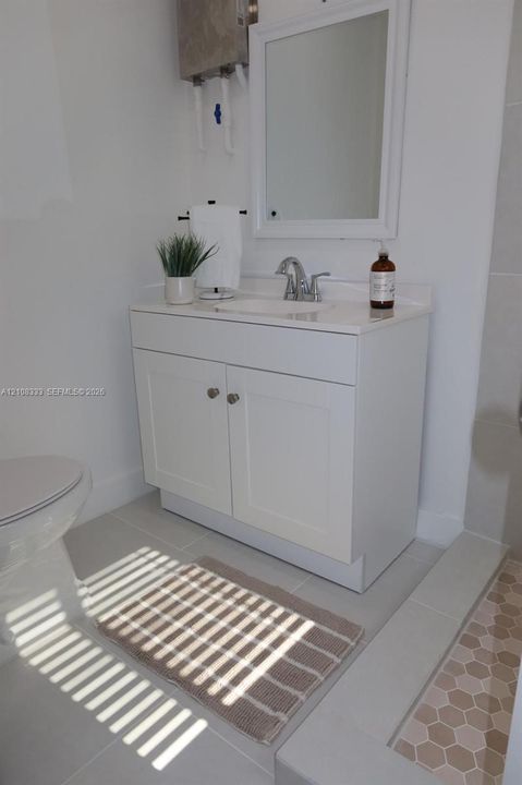 White shaker-style vanity with a solid surface countertop, integrated sink, chrome faucet, wall-mounted mirror, and hexagon-patterned floor tile