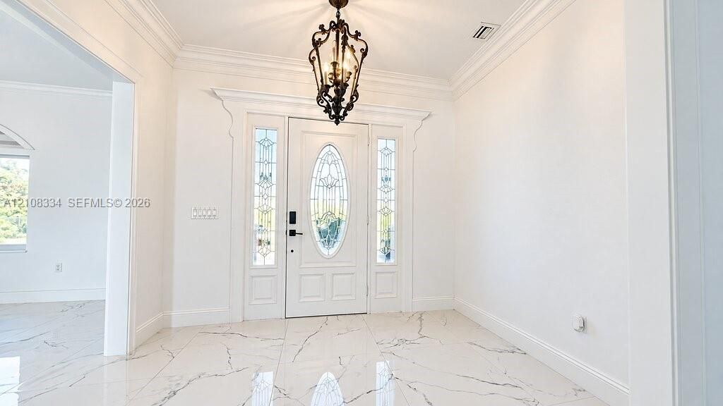 Foyer featuring large-format tile flooring with marble-style veining, crown molding, iron lantern-style pendant lighting, and a white front entry door with a decorative oval glass inlay flanked by sidelights