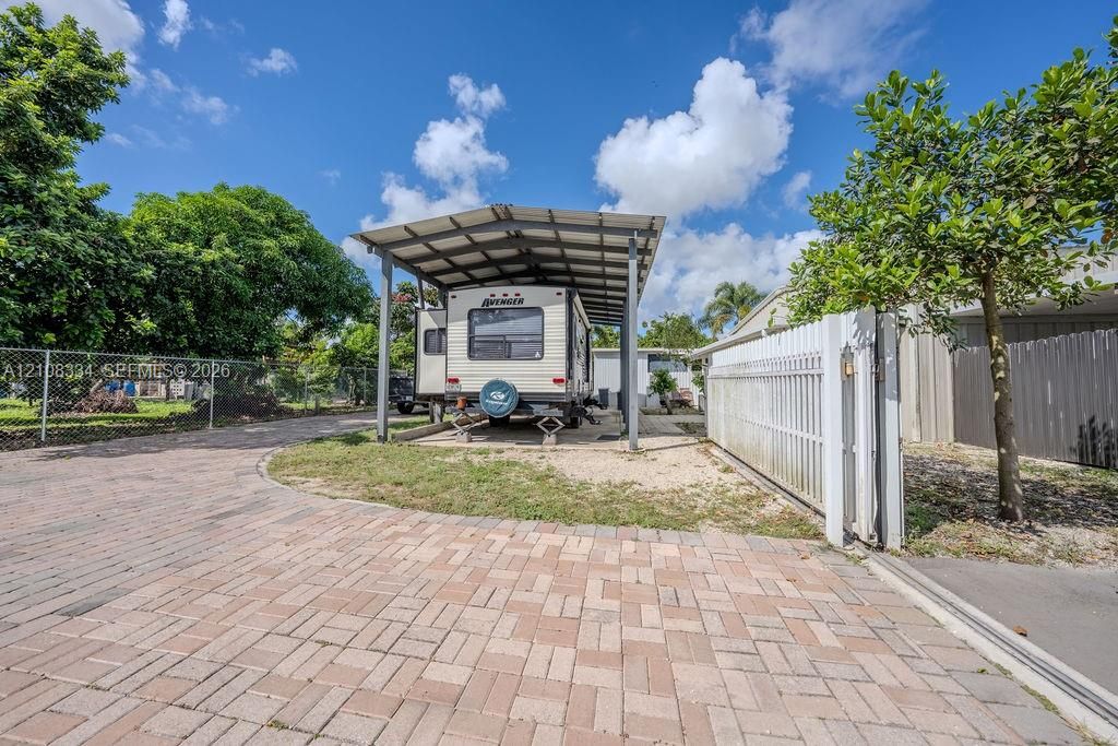 Paver driveway featuring a metal-frame covered carport, perimeter chain-link fencing, and white vertical-slat side gate