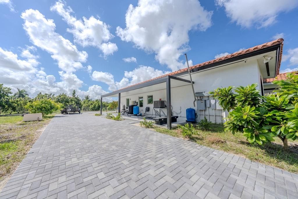 Stucco exterior with a terracotta-style tile roof, expansive gray paver driveway, and a black metal-frame covered side patio