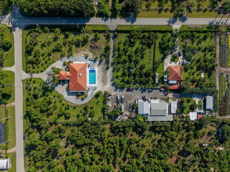 Aerial perspective of a sprawling property featuring multiple structures, a rectangular swimming pool, expansive grounds with mature landscaping, and an extensive driveway system