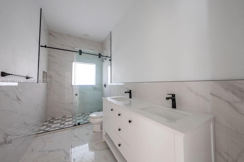 Dual-sink vanity with a white countertop and matte black fixtures, walk-in shower with a glass sliding door, large-format stone-patterned tile flooring, and floor-to-ceiling tiled shower surround