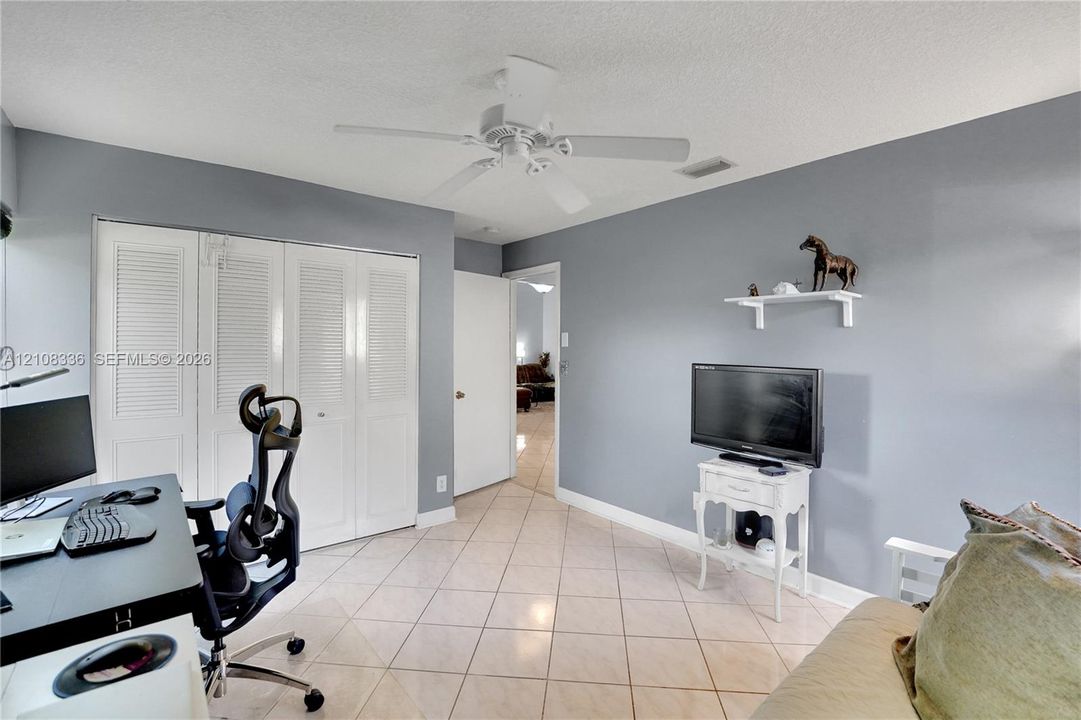 Room featuring neutral-toned walls, tile flooring, louvered bi-fold closet doors, a white ceiling fan, and a wall-mounted display shelf