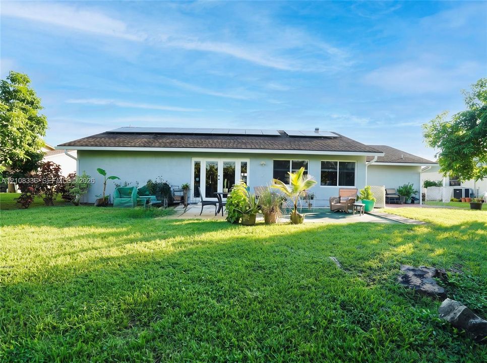 Exterior rear elevation featuring a stucco facade, rooftop solar panel array, multi-panel glass door, and a paved patio area set within a spacious lawn