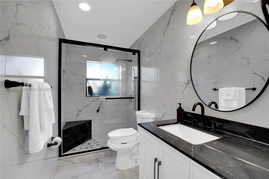 Bathroom featuring large-format wall tiling, a walk-in glass-enclosed shower with a built-in bench, a custom vanity with a contrasting dark stone countertop, a circular wall-mounted mirror, and matte black hardware
