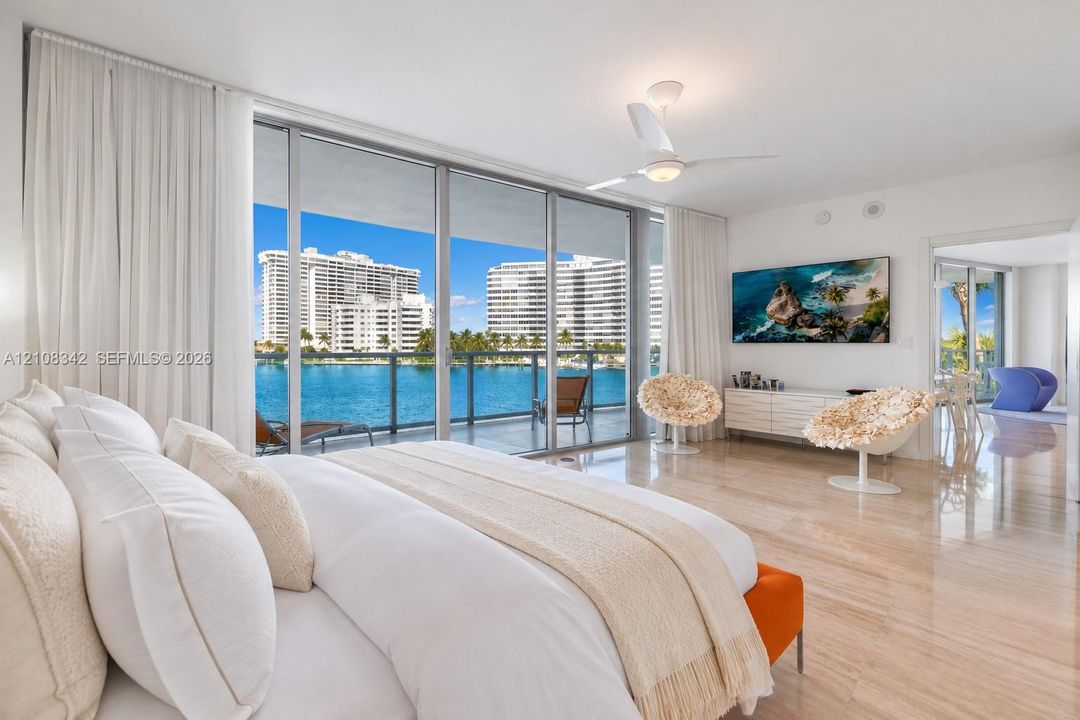 Floor-to-ceiling sliding glass doors, polished stone flooring, private glass-railed balcony, built-in ceiling fan, and prominent water backdrop