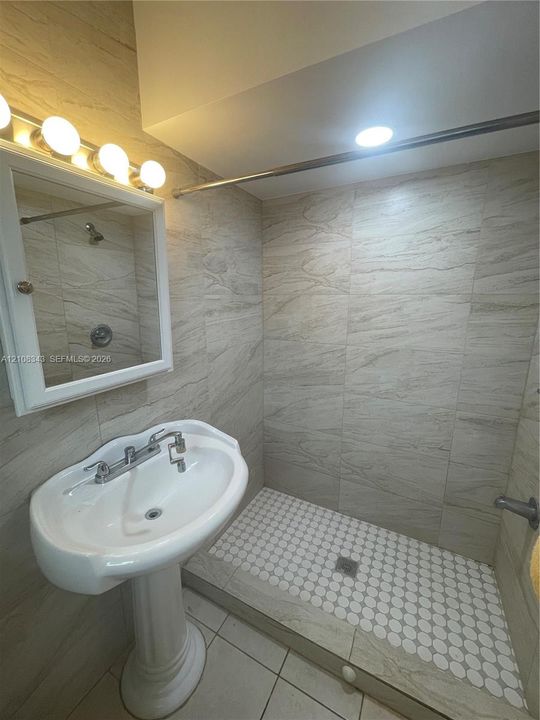 Bathroom featuring floor-to-ceiling neutral stone-finish tile, walk-in shower with penny-round floor tile, pedestal sink with chrome faucet, and mirrored medicine cabinet with integrated four-light fixture