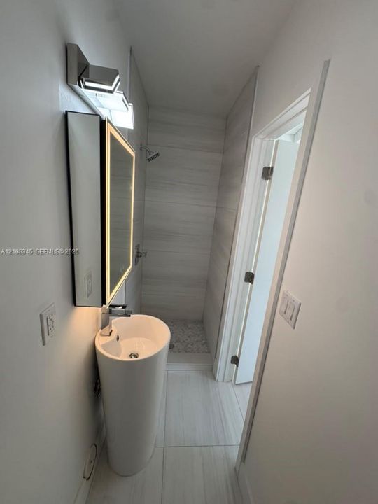 Pedestal sink featuring a chrome faucet, illuminated vanity mirror, shower with light-toned wall tile, and textured stone shower floor tiling