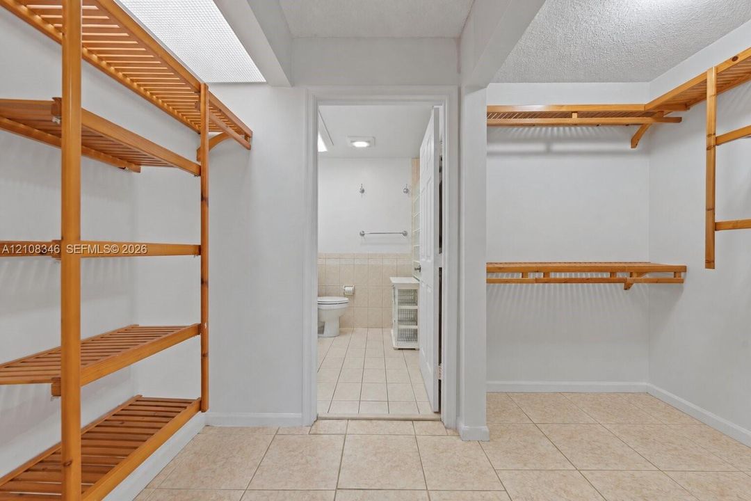 Walk-in closet featuring light-toned tile flooring and custom wood slatted shelving, with entry to a bathroom showcasing tile wainscoting and a white vanity cabinet