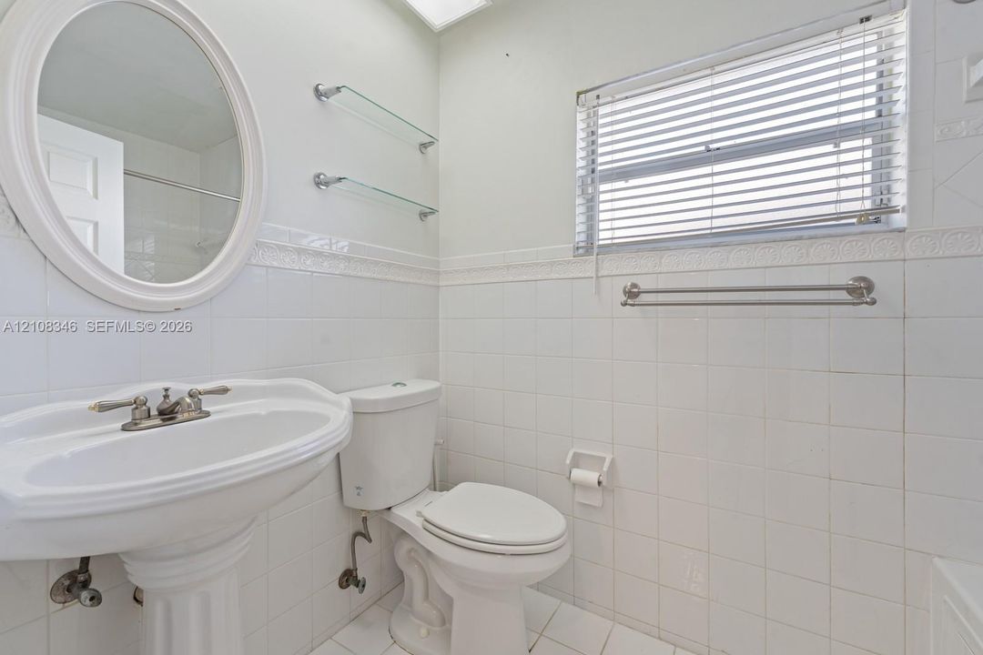 Bathroom featuring white ceramic tile wall cladding, pedestal sink with polished metal faucet, oval wall-mounted mirror, glass shelving, and horizontal window blinds