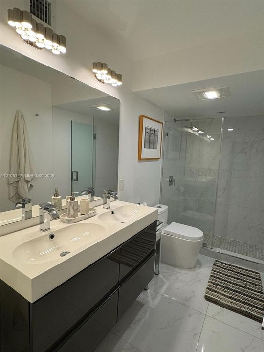 Dual-sink vanity with high-gloss cabinetry and integrated countertop, glass-enclosed walk-in shower with tiled surround, and polished large-format floor tiling