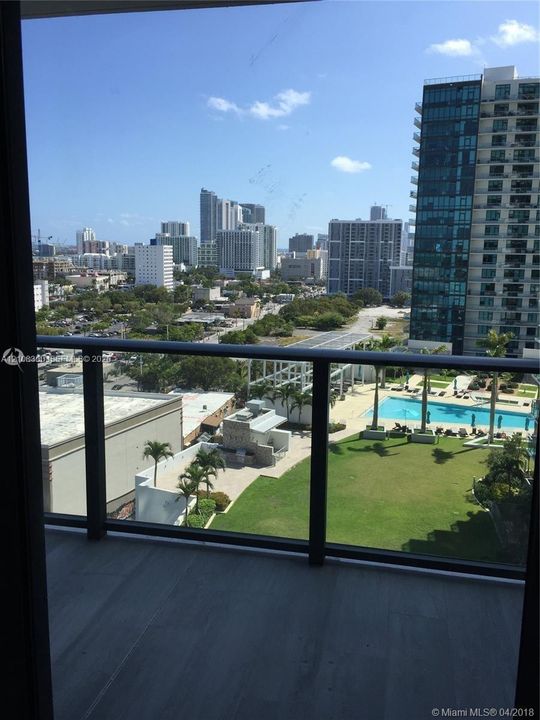 Private balcony featuring metal railings and a glass panel surround, overlooking an outdoor swimming pool, grass lawn, and urban cityscape