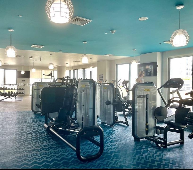 Fitness center featuring floor-to-ceiling windows, blue chevron-patterned flooring, suspended globe pendant lighting, and multiple wall-mounted televisions