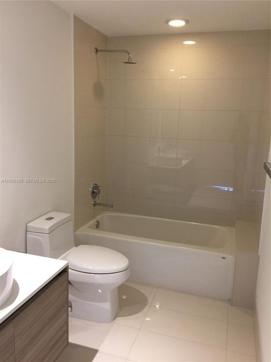 Full-height neutral wall tiling, bathtub with integrated shower, vanity with wood-finish cabinetry, white countertop, and recessed ceiling lighting