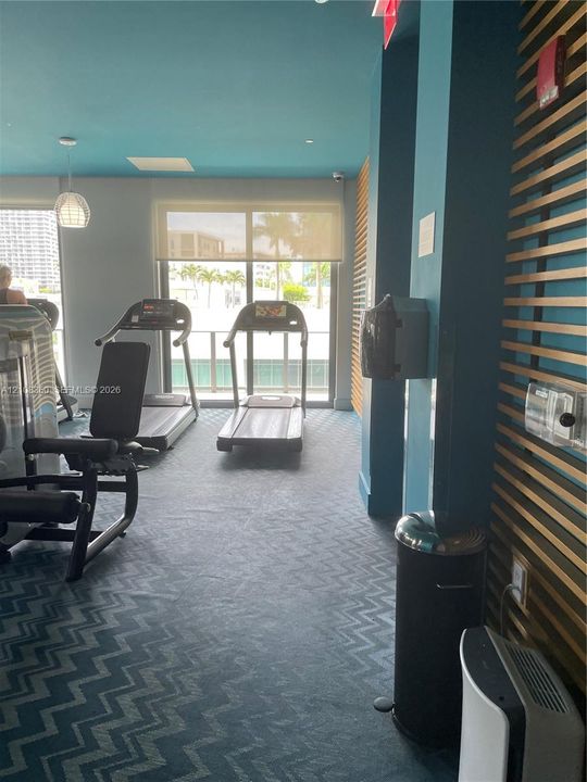 Fitness center featuring chevron-patterned carpeting, decorative wood-slat wall paneling, and floor-to-ceiling glass sliding doors with solar shades