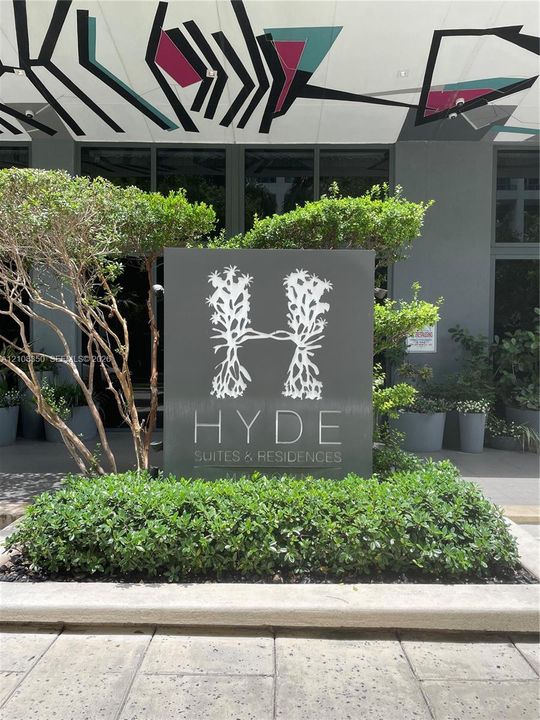 Property entrance featuring custom signage, concrete masonry, manicured shrubbery, mature landscaping, and structural ceiling art