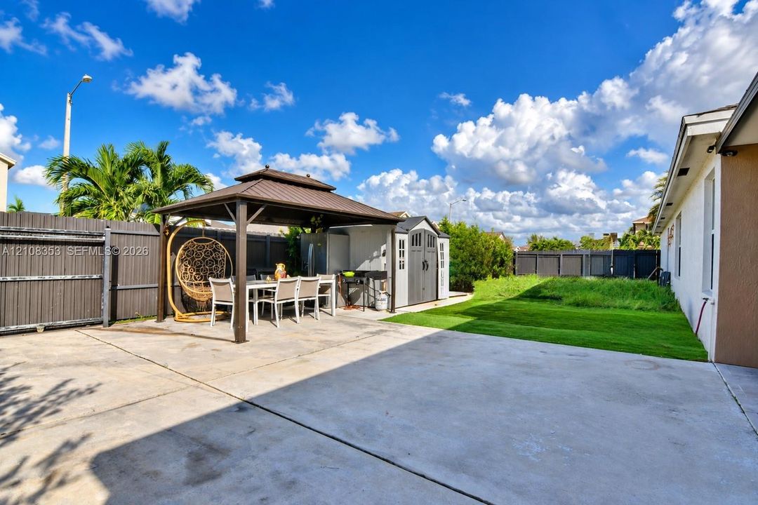 Spacious concrete patio area featuring a metal-frame gazebo, synthetic turf lawn, privacy fencing, and an exterior storage structure
