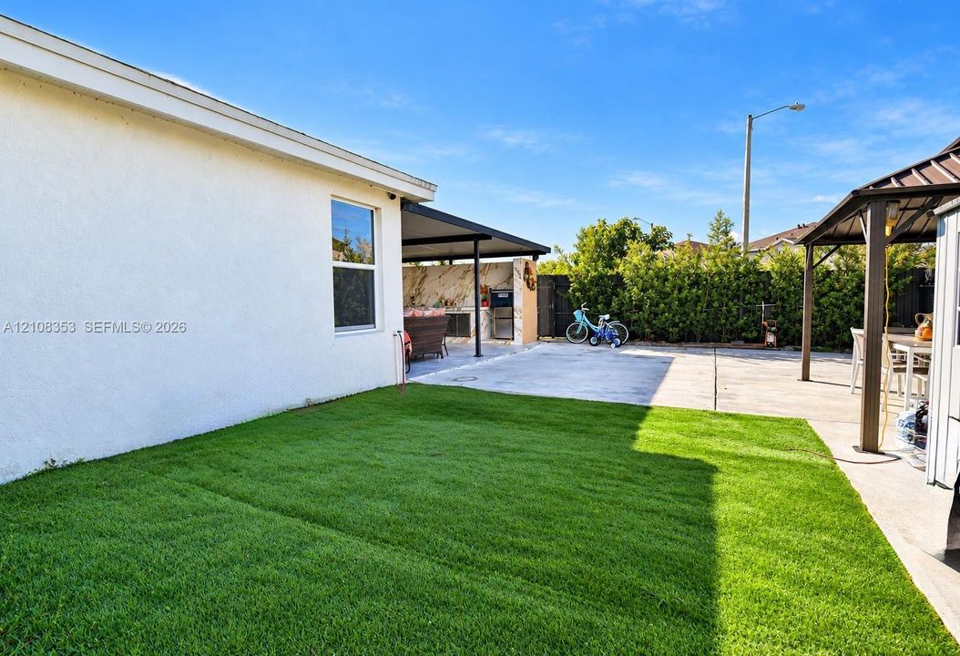 Backyard featuring a concrete patio, a covered outdoor kitchen area, a permanent metal-framed gazebo, and a manicured lawn