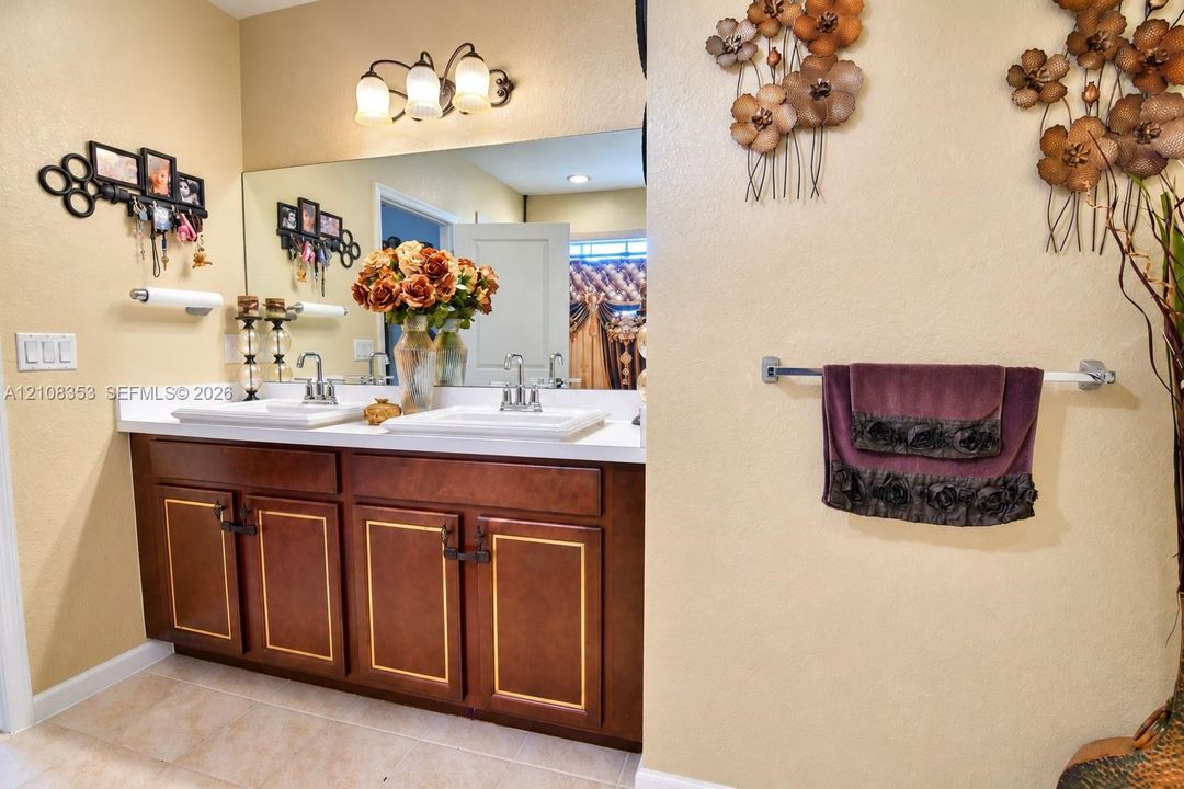 Dual-sink vanity with white surface, wood cabinetry featuring gold-toned trim, wall-mounted three-light fixture, and large rectangular mirror