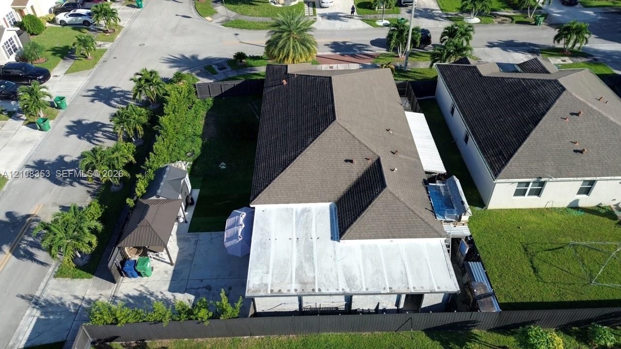 Aerial perspective featuring a multi-pitched shingle roof, a covered patio structure, a paved side area, and perimeter fencing