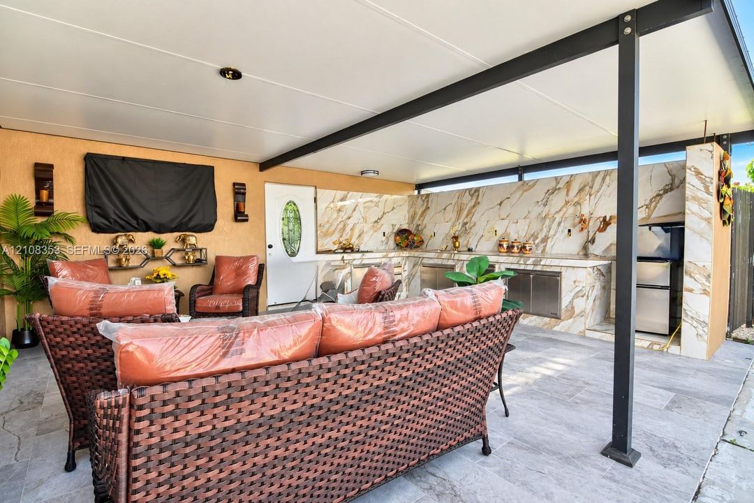 Covered outdoor patio featuring large-format tile flooring, a built-in outdoor kitchen with stone-patterned backsplash, stainless steel appliances, and a structural metal support frame