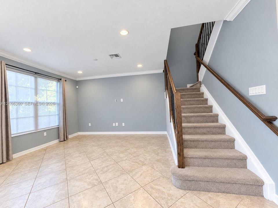 Open interior space featuring neutral tile flooring, recessed lighting, crown molding, and a staircase with a wooden handrail and metal balusters