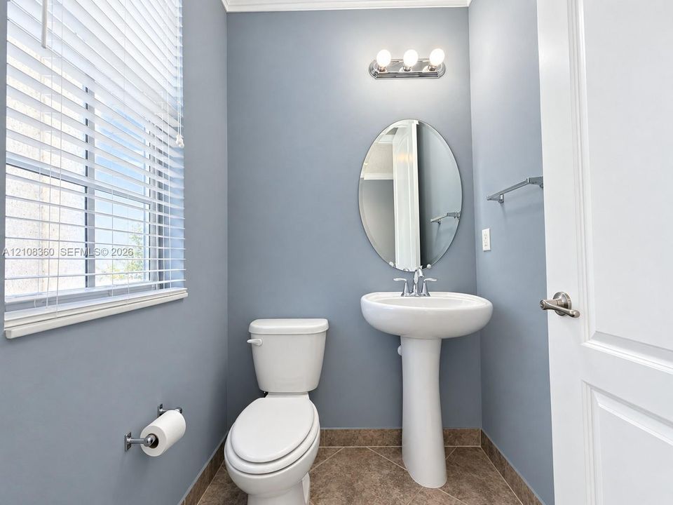 White pedestal sink with chrome faucet, oval wall-mounted mirror, three-light vanity fixture, and tile flooring