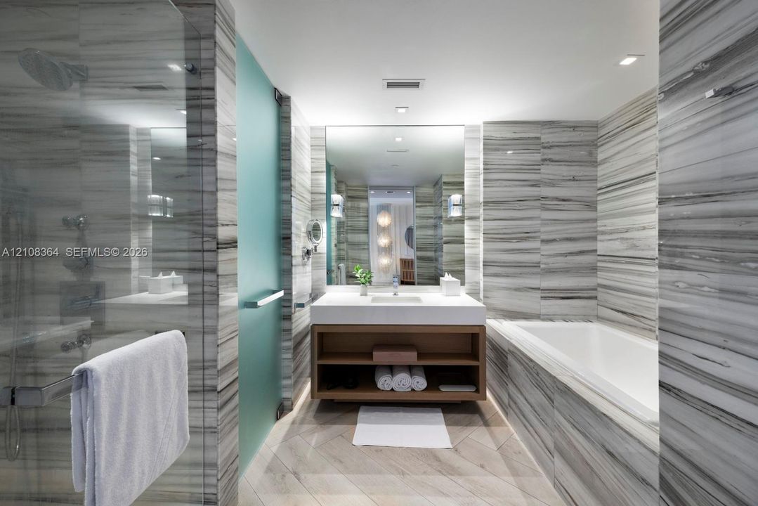 Bathroom featuring floor-to-ceiling stone tile, a soaking tub, a glass-enclosed shower, a floating wood-finish vanity, and recessed lighting