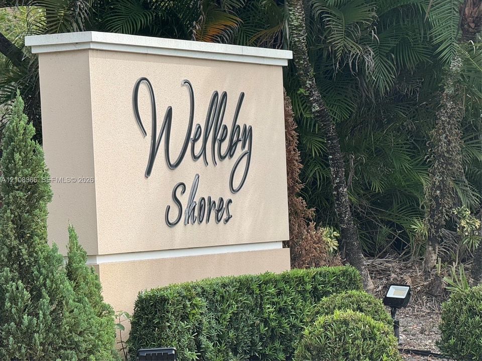 Stucco monument sign featuring dimensional metal lettering, capped with a stone cornice, and accented by manicured landscape hedging and mature palm trees