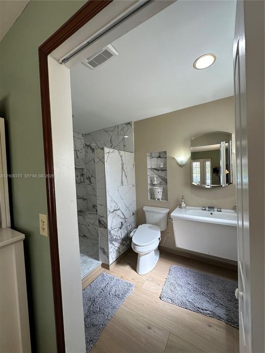 Walk-in shower with floor-to-ceiling veined tile surround, recessed wall shelving, wall-mounted vanity, and wood-finish flooring
