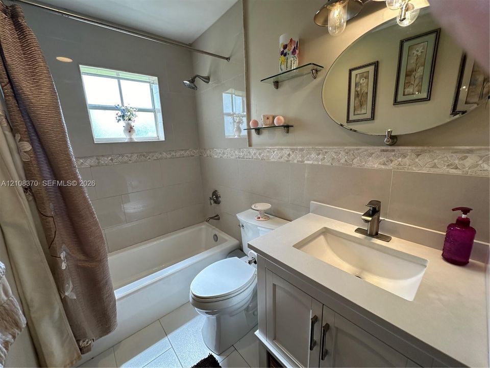 Bathroom featuring a vanity with a solid-surface countertop, an integrated sink, an oval wall-mounted mirror, neutral tile wall surround with decorative accent inlay, and a tub-shower combination