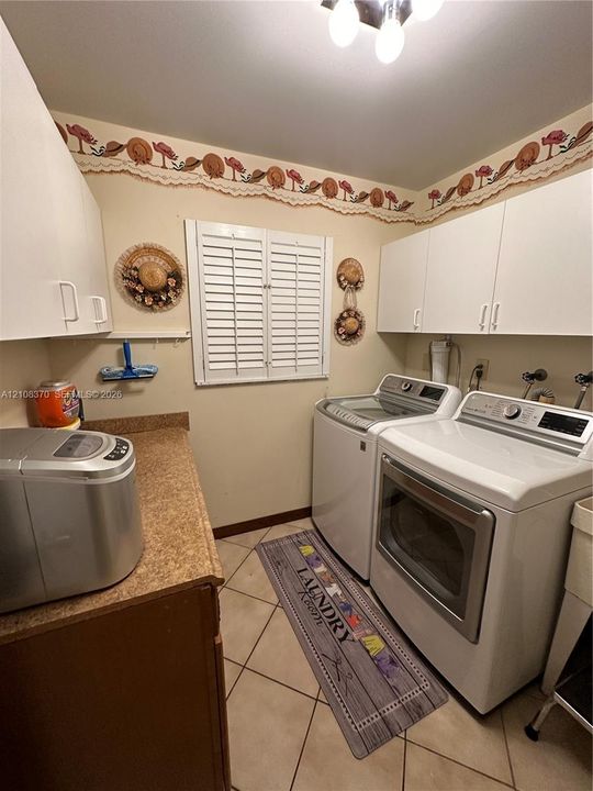 Laundry room featuring white shaker-style wall cabinetry, a laminate countertop, tile flooring, and a window with plantation-style shutters