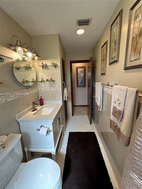 White vanity with integrated basin, polished metal hardware, circular wall-mounted mirror, glass shelving, and tile wainscoting