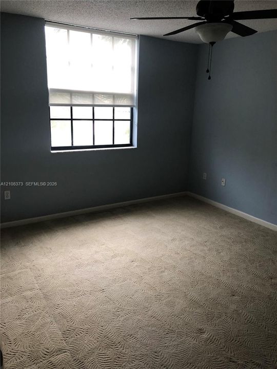 Room featuring patterned carpeting, a multi-pane window with a roller shade, textured ceiling, and a ceiling fan with integrated light fixture