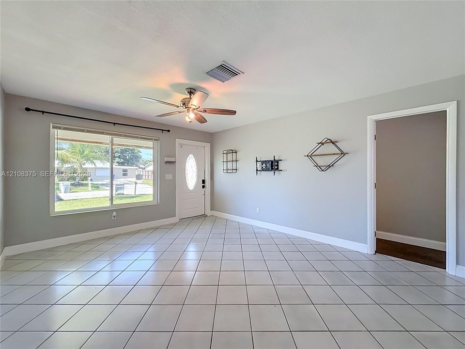 Neutral-toned interior featuring large-format ceramic tile flooring, a multi-pane window with hardware, a solid-panel front door with an oval glass insert, and a ceiling-mounted fan with integrated lighting