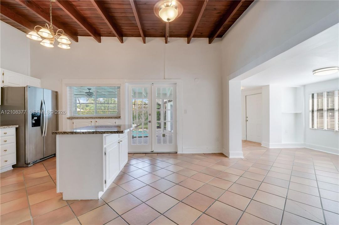 Exposed wood beam ceiling, tile flooring, kitchen island with granite countertop, stainless steel appliances, and french double doors leading to the patio and pool area.