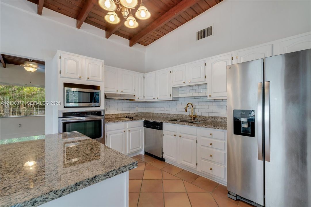 Another view of the Kitchen featuring white cabinetry, stone countertops, stainless steel appliances, a white tile backsplash, and a vaulted ceiling with exposed wood beams