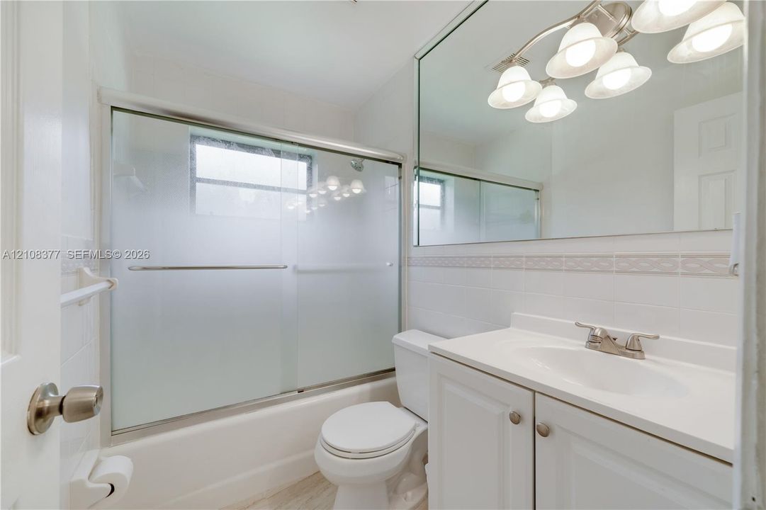 Hall bathroom featuring a tub-and-shower combination with a sliding glass door, white cabinetry with a solid surface countertop, an integrated basin, a large wall-mounted mirror, and multi-bulb overhead lighting