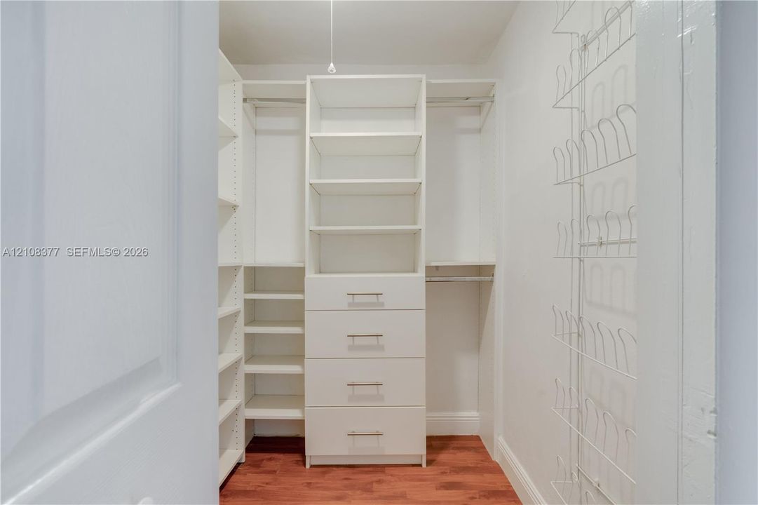 Walk-in closet featuring white built-in shelving, a central stack of drawers with metal pulls, dual hanging rods, and wood-finish flooring