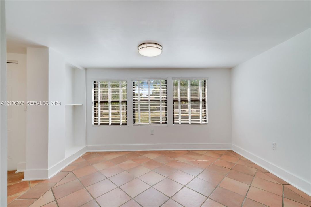 Terra-cotta tile flooring, three large windows with horizontal blinds, built-in recessed shelving, and a flush-mount circular ceiling light