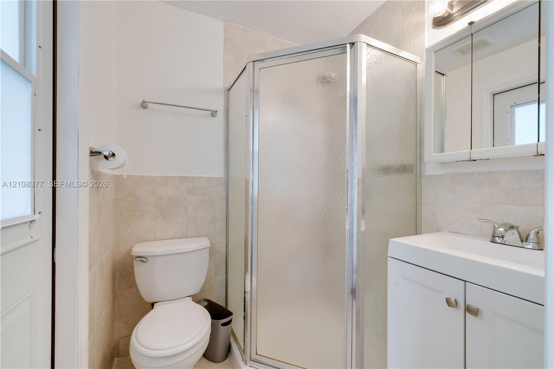 Corner shower enclosure with textured glass doors, vanity cabinet with integrated sink, dual-door mirrored medicine cabinet, and tiled wainscoting