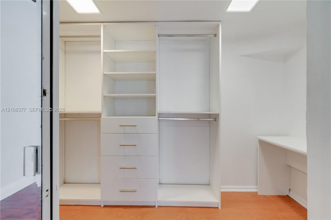 Custom closet system featuring white cabinetry with multiple hanging rods, open shelving, four storage drawers with metallic hardware, and a built-in workstation surface