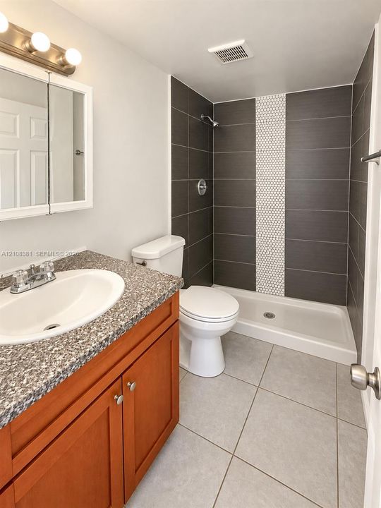 Bathroom featuring a vanity with speckled countertop and integrated oval sink, wood-finish cabinetry, shower stall with large-format dark tile and a central vertical mosaic accent, and neutral floor tile