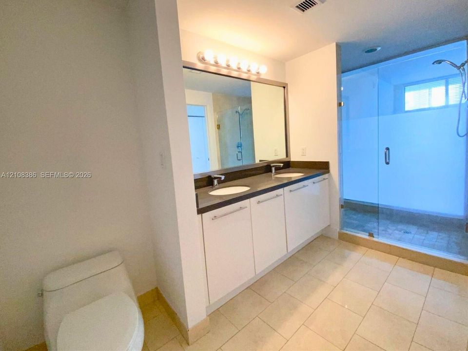 Bathroom featuring a dual-sink vanity with white cabinetry, dark stone-composite countertop, full-width mirror, and a glass-enclosed walk-in shower with tile flooring