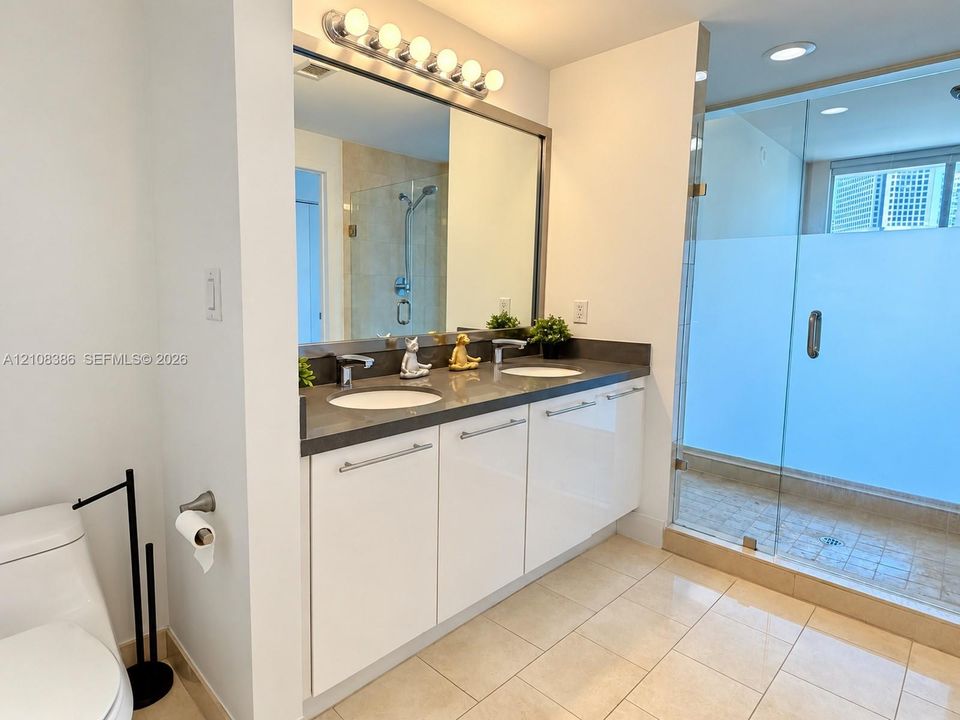 Double vanity with high-gloss cabinetry, dark solid-surface countertops, dual undermount sinks, frameless glass-enclosed shower, and tile flooring