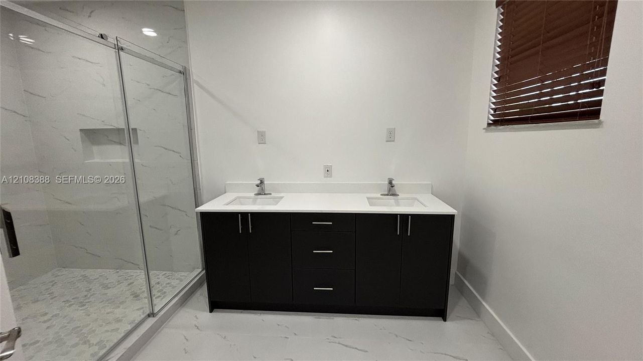 Double vanity with dark cabinetry, white countertop, dual undermount sinks, and a glass-enclosed shower with tiled walls and built-in niche