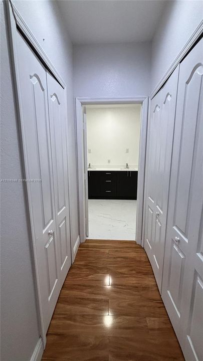 Hallway featuring dual white bi-fold closet doors, wood-finish flooring, and a stone-topped dark-finish vanity visible through the doorway