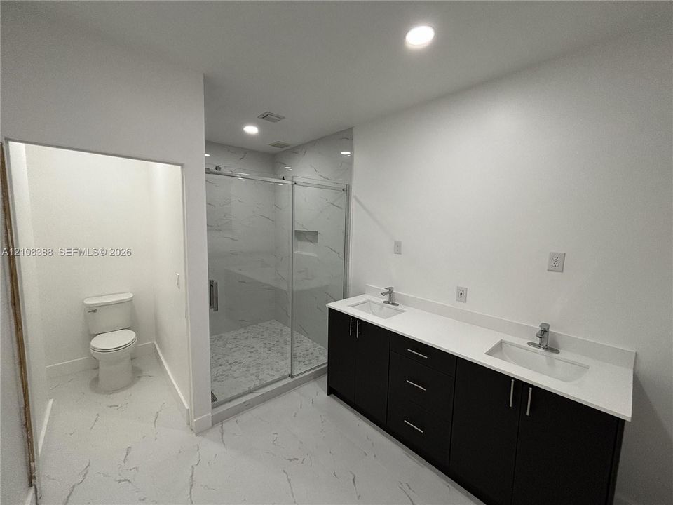 Dual-sink vanity with dark cabinetry and stone-finish countertop, walk-in shower with glass enclosure, and light-toned tile flooring
