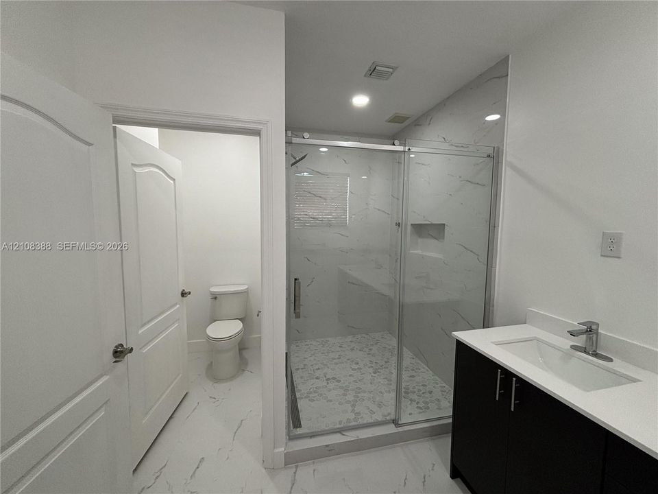 Bathroom featuring a dark wood-finish vanity with a white countertop, a walk-in shower with stone-patterned tile walls and a pebble-stone floor, and a separate water closet area